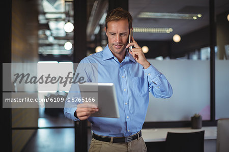 Businessman looking at digital tablet while talking on mobile phone in the office