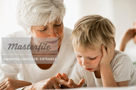 Mature woman and grandson lie on their fronts on a bed side by side while looking down.