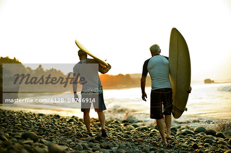 Two men walk along a beach carrying a surfboard each as the sun rises ahead of them.