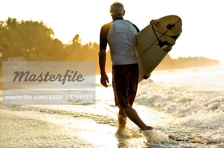 Man walks through the surf carrying a surfboard under his arm as the sun rises ahead of him.