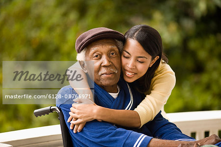 Young woman leans over and embraces her grandfather sitting in a wheelchair as they pose for a portrait on a porch.