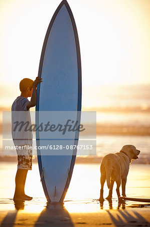 Teenage boy standing with a surfboard on a beach at sunset.