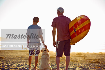 Senior man standing on a beach with his grandson and a surfboard.