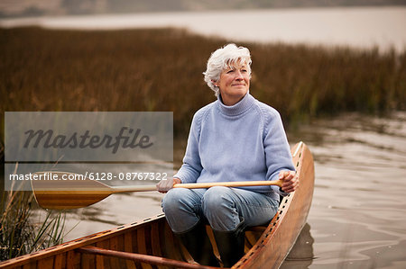 Mature woman smiles and looks away as she holds an oar ready to row and sits in the aft of a wooden canoe on a lake.