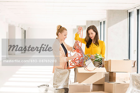 Young businesswomen untangling cords while standing by cardboard boxes in new office
