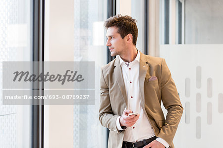 Young businessman holding mobile phone while looking through office window