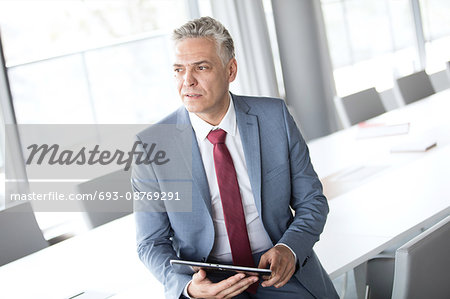 Thoughtful mature businessman holding digital tablet while leaning on conference table