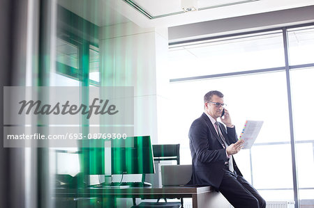 Mature businessman holding document while talking on mobile phone in office