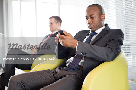 Young businessman using mobile phone on chair with male colleague in background at office