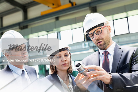 Mid adult businessman explaining machine part of colleagues in metal industry