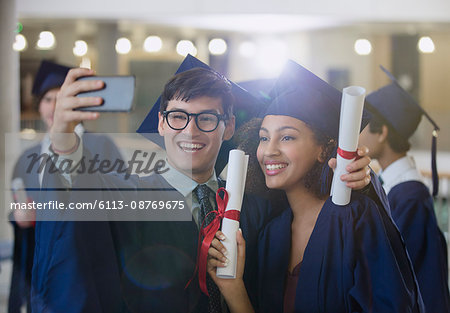College graduates in cap and gown holding diplomas posing for selfie