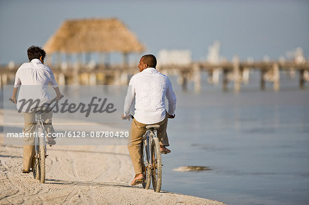 Mid-adult man and his son riding bicycles on a sandy beach near the water.