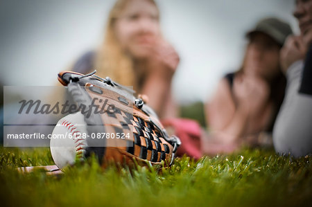 Baseball lying inside a baseball mitt on grass in front of a group of young people.