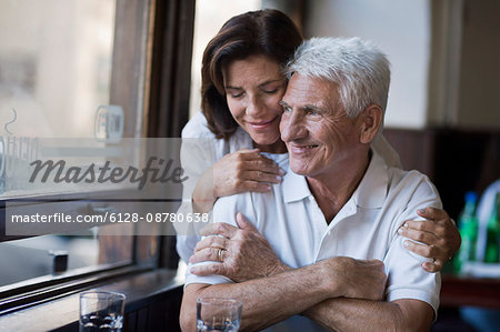 Senior woman embraces her husband as he looks out the window of a cafe.