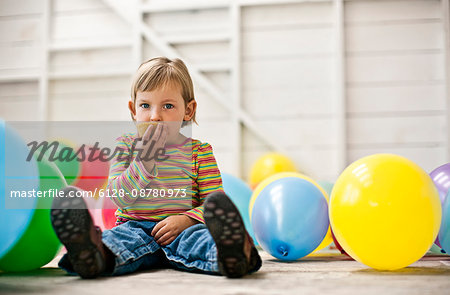 Little girl sitting in a room full of balloons and eating a cupcake.