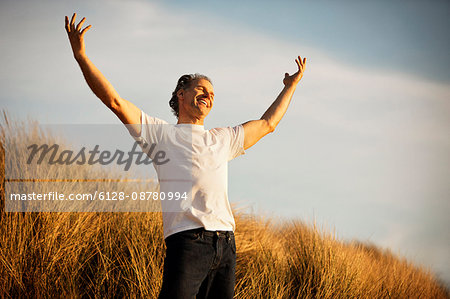 Happy mature man standing on a grassy sand dune with his arms raised.