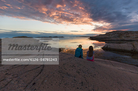 Sweden, Gotaland, Bohuslan, Grebbestad, Tjurpannan, Girls (6-7) sitting on rocky seashore at sunset