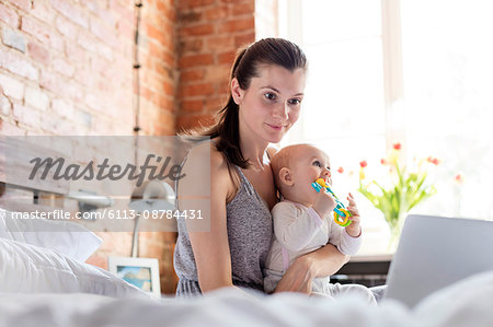 Mother holding daughter and working at laptop on bed