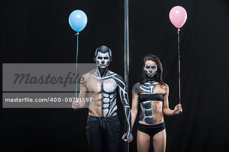 Gorgeous couple of pole dancers holds their hands together next to a pylon on a dark background in the studio. They have body-art and wears black clothes. Girl and guy hold blue and pink balloons.