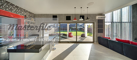 Interior in a modern style with a kitchen zone with a bar, concrete and brick walls, large windows and a door. On the right there is dark sofa with red pillows. Outside there is a terrace and a lawn.