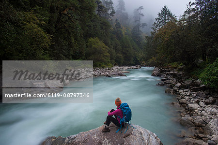 A woman takes a break from the trail and sits beside the Langtang Khola near the little village of Riverside on a misty evening, Nepal, Himalayas, Asia