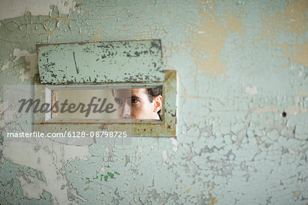 Mid-adult businessman peering through a hole in a wall of a derelict building.