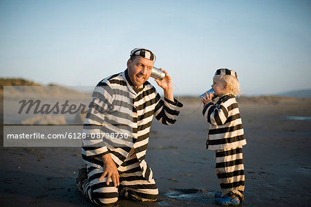 Mid-adult father and his son playing with a tin can phone on a beach while wearing striped prisoner costumes.