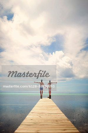 Young couple hold hands with arms outstretched, while balancing on an ocean pier.