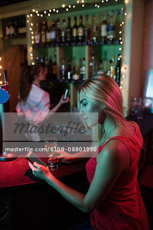 Woman using mobile phone while having a glass of wine at counter in bar