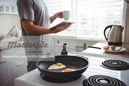 Man using digital tablet while preparing fried eggs in the kitchen at home