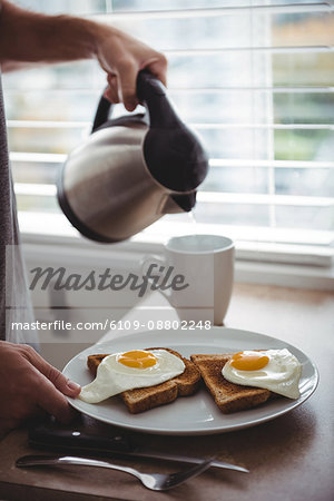 Man holding his breakfast plate while pouring hot water into mug in the kitchen