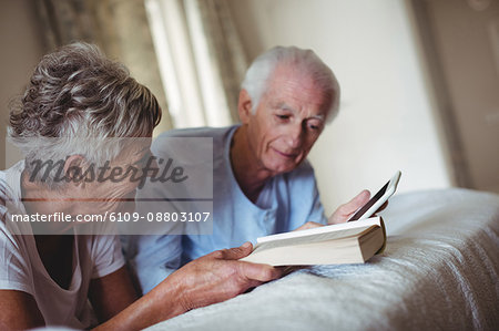 Senior couple using digital tablet and reading book on bed in bedroom