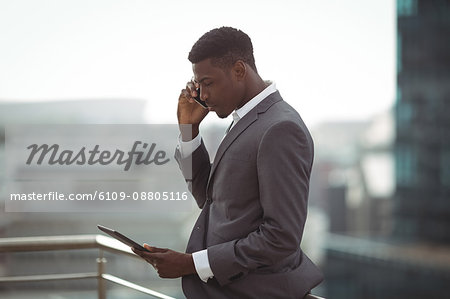 Businessman using digital tablet and talking on mobile phone on office terrace