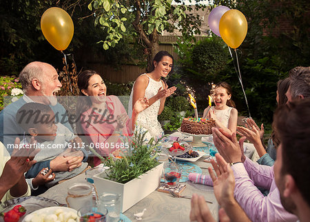 Multi-ethnic multi-generation family clapping celebrating birthday with fireworks cake at patio table