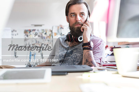 Pensive design professional with headphones talking on cell phone at desk