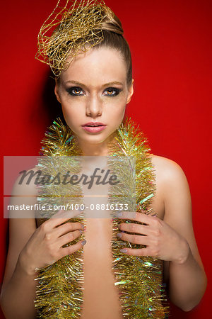 stunning woman with charming expression posing on red background with glossy golden make-up and sparkly hair-style accessory and tinsel on her nude breast.