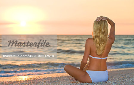 A relaxed sexy young blonde woman or girl wearing a bikini sitting on a deserted tropical beach at sunset or sunrise