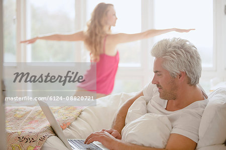 Woman practicing yoga warrior 2 pose in bedroom with husband using laptop in bed