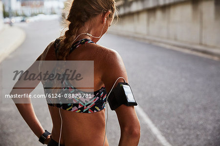 Fit female runner in sports bra with mp3 player armband and headphones resting on urban street