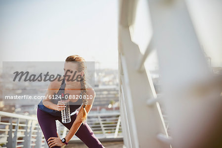 Female runner resting stretching leg and drinking water on sunny urban footbridge