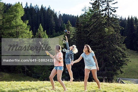 Three female adult friends blowing and jumping for bubbles in field, Sattelbergalm, Tirol, Austria