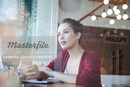 Young woman sitting in cafée, holding smartphone, looking out of window