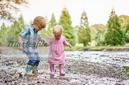 Brother and sister holding hands splashing in muddy puddle
