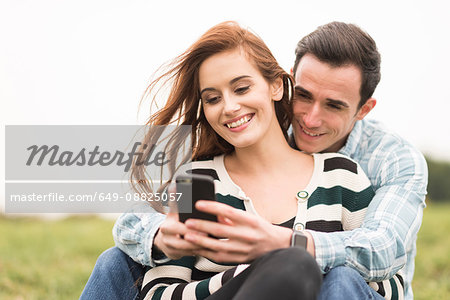 Couple sitting in field hugging looking at smartphone