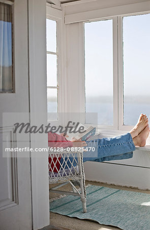 Man sitting with a book in a wicker chair inside a sunny room.