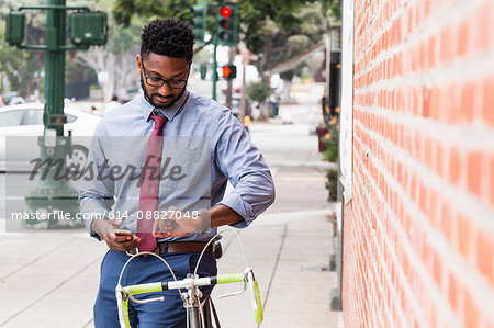 Young man standing beside bicycle, using smartphone, looking at wristwatch