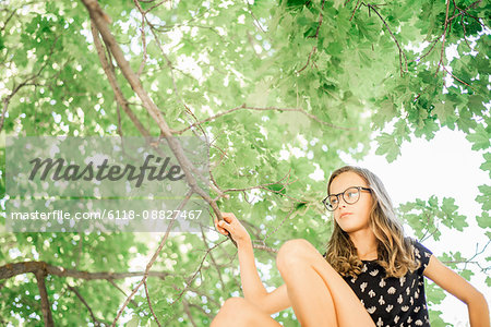 A young girl in shorts sitting on a high tree branch under a canopy of green leaves.