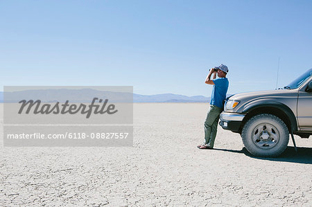 Man standing on vast desert, looking through binoculars and leaning against truck, Black Rock Desert