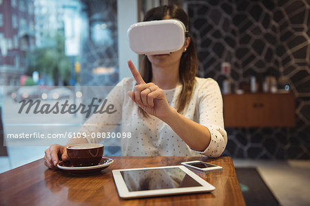 Businesswoman using vr headset while sitting at table with coffee, digital tablet and phone
