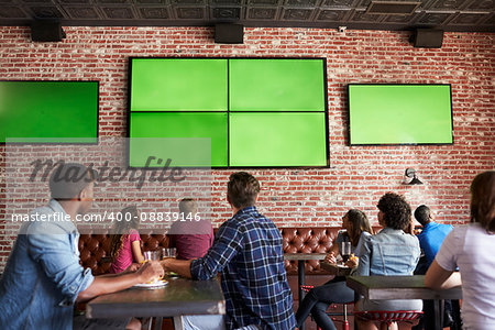 Rear View Of Friends Watching Game In Sports Bar On Screens